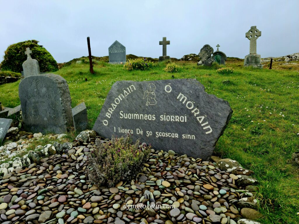 Gravestone with writing in Gaelic - O'Braonain on one side and O' Moráin on the other with an angel etched between them and underneath: suaimhneas síorraí I líonta Dé go gcastar sinn. The ground in front is full of pebbles and on the horizon in the background are a line of crosses and other headstones