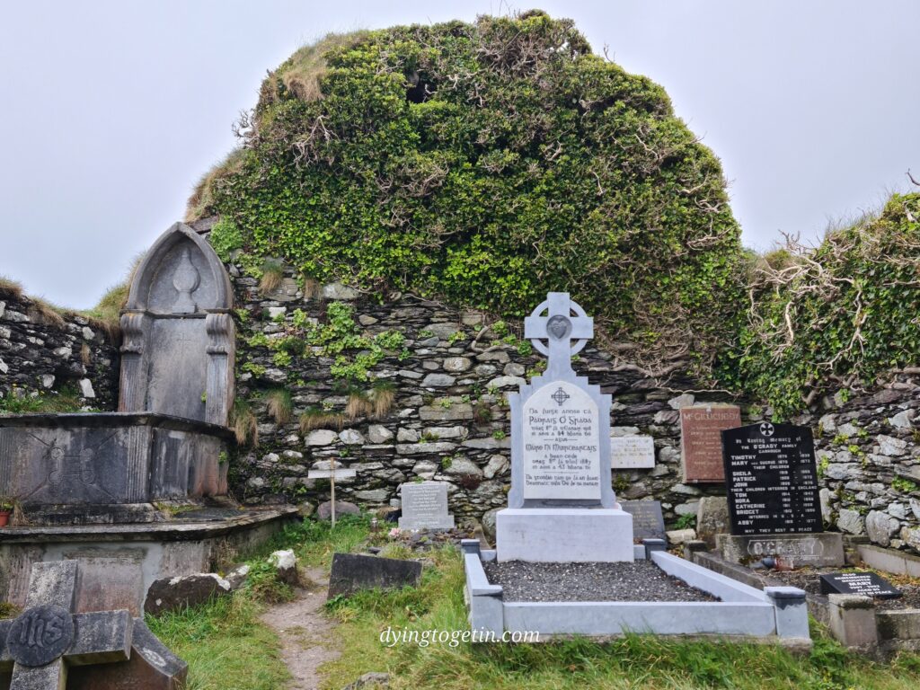 Interior of a ruined, roofless church, the back wall covered in ivy. A tombstone is tucked into the left corder, and other gravestone are visible too