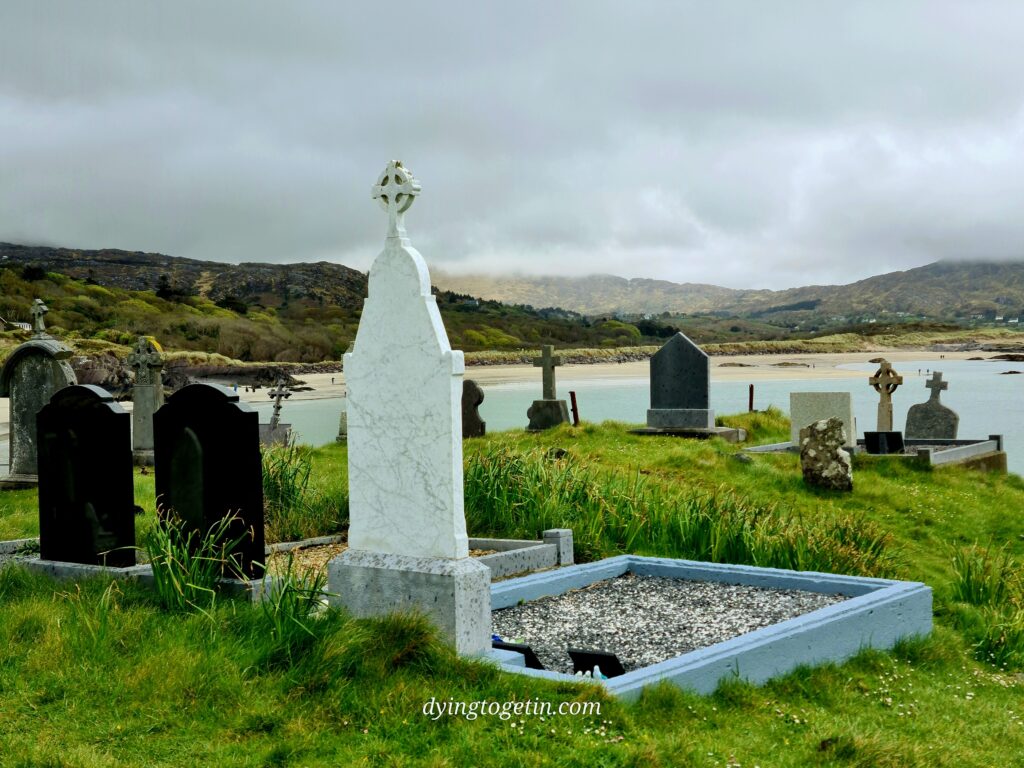 The backs of headstones on a hill facing out over the ocean. In the nearground is a tall white slab with a celtic cross head on top. The grave surround is blue stone. 