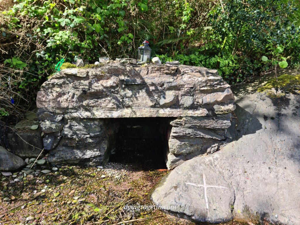 Stone wellhouse built over a trickle of water. To the side is a stone with a crude white cross etched into it. 