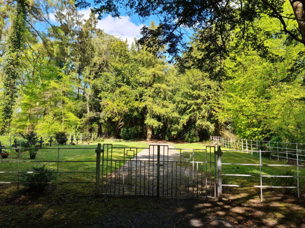 Metal gates leading to a walk way flanked by black crosses on the left with tree in full leaf in the distance