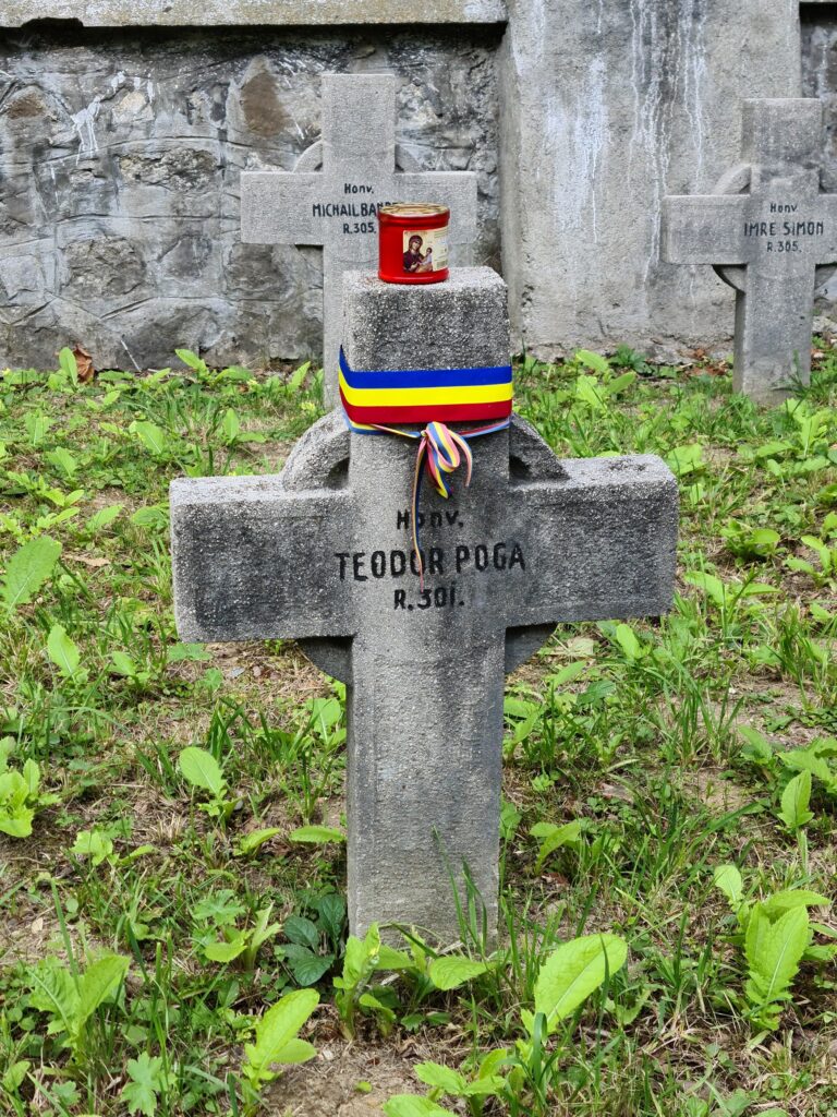 Stone cross with blue, yellow, red ribbon around the top. A red candle holder sits on top. Inscription reads Honv. Teodor Poga. R. 501. Two other crosses in the background, ribbonless, inscribed Michail B,,,, Imre Simon