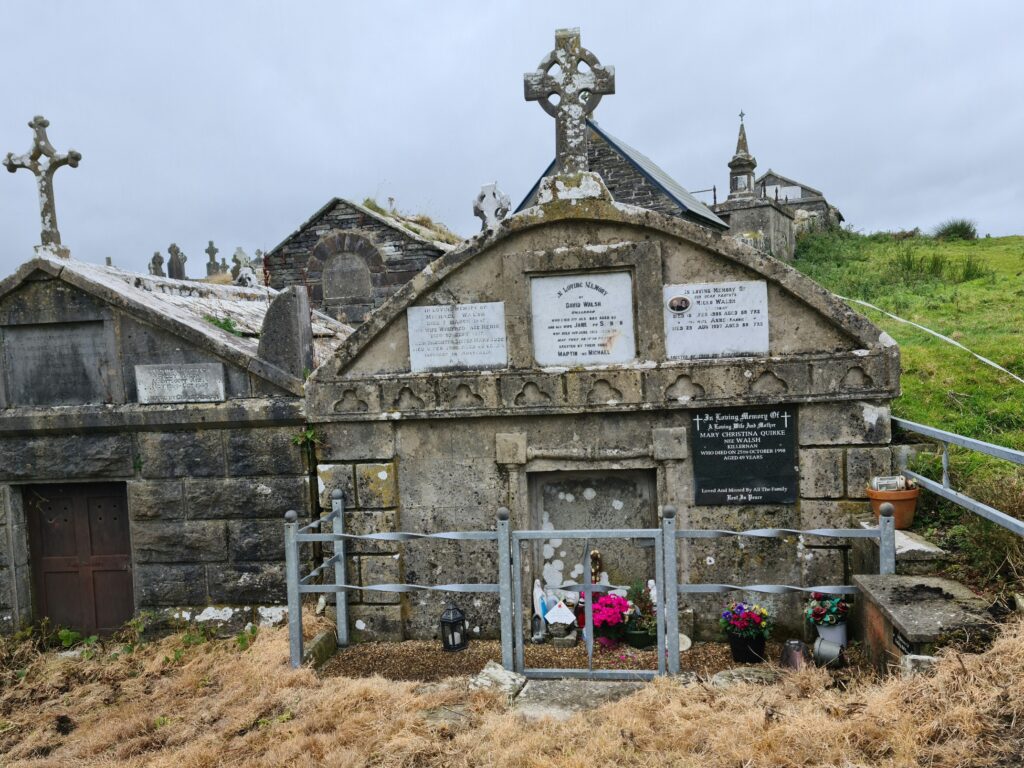 Gabled tombs one with three plaques on the wall, a grey iron fence around it, and some pink flowers in the foreground. The apex of the roof bears a celtic cross. 