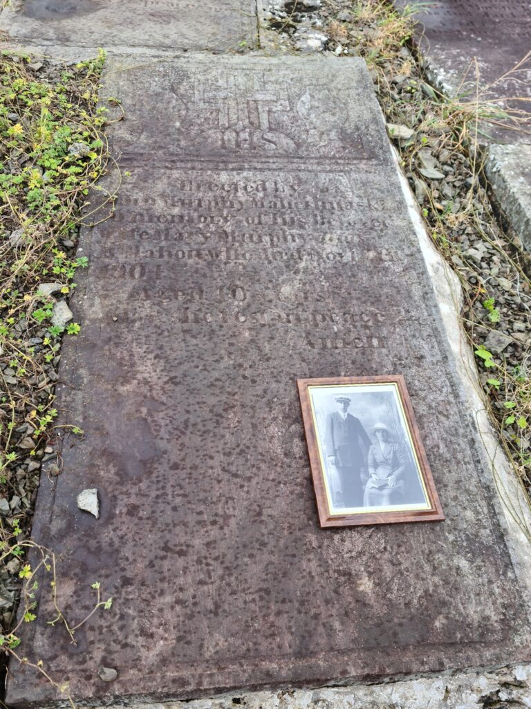 Old gravestone set in the ground with faded inscription. ON it sits a framed black-and-white photo of a standing man and a seated woman. 