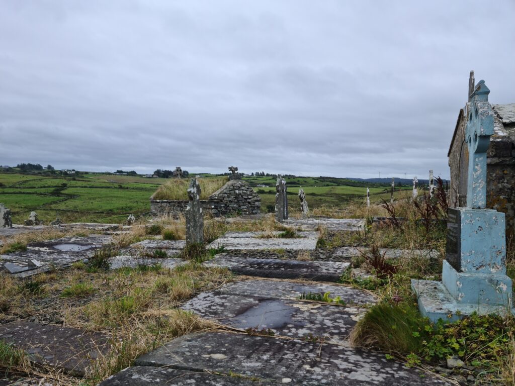 Old graveyard with gravestones and Celtic crosses (one in a pale blue to the right) loverlooking farmed fields. 