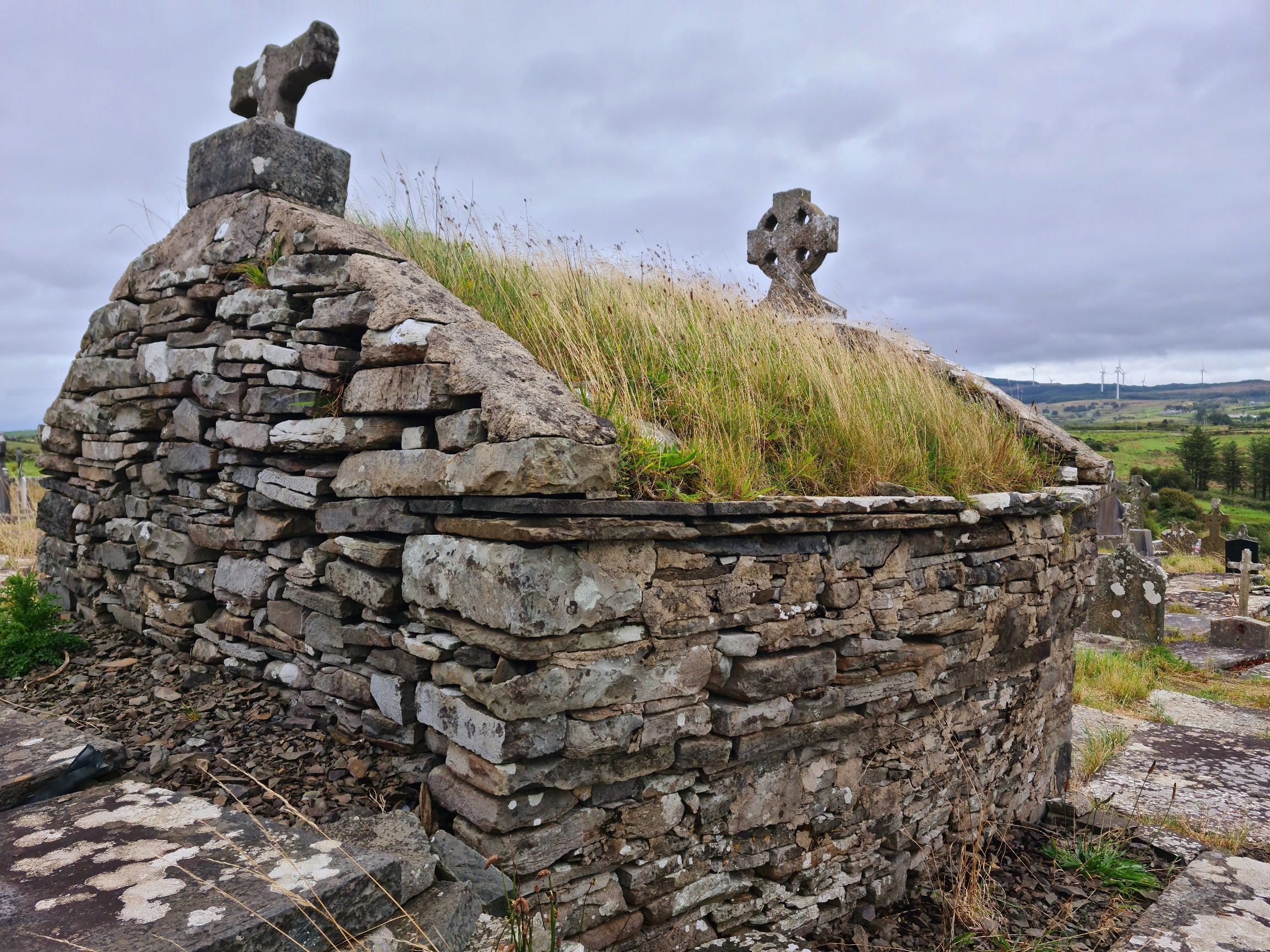 Small stone building with a pitched roof covered in grass. A stone cross sits on the apex on the right and a broken one on the left.