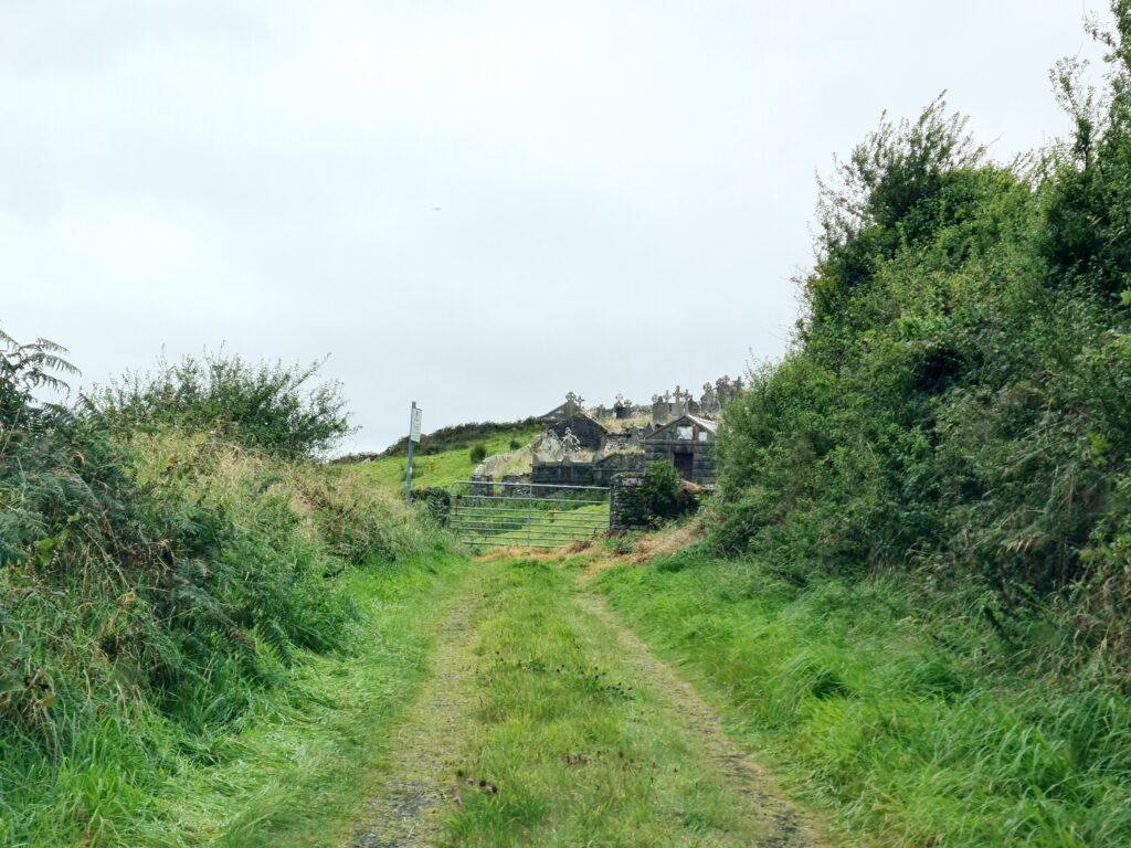 Boreen road, the width of one car, with a grass meridian leads to a gate behind which there are gravestones and tombs