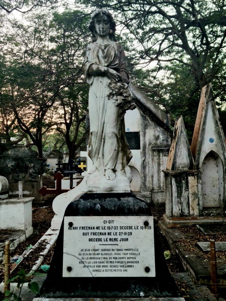 Gravestone with a statue of a young woman - possibly an angel - with a bunch of flowers. The inscription is in French: CI-GIT ENRI FREEMAN NE LE 15-7-32 DECEDE LE 10-9-38 GUY FREEMAN NE LE 27-11-39 DECEDE LE MENE JOUR JE SUIS VIVANT QUOIQUE MA TOMBE IMMORTELLE AU SEIN DU CHAMP GLORIEUX IMAGE TROP CRUELLE OU LE BERCEAU FINAL DE MON CORPS DEPOSE RESTE LE LIEU SAINT DE MES PARENTS SACRIFIES JEVEILLE SUR VOUS SECHEZ VOS LARMES SONGEZ A CETTE PETITE AME