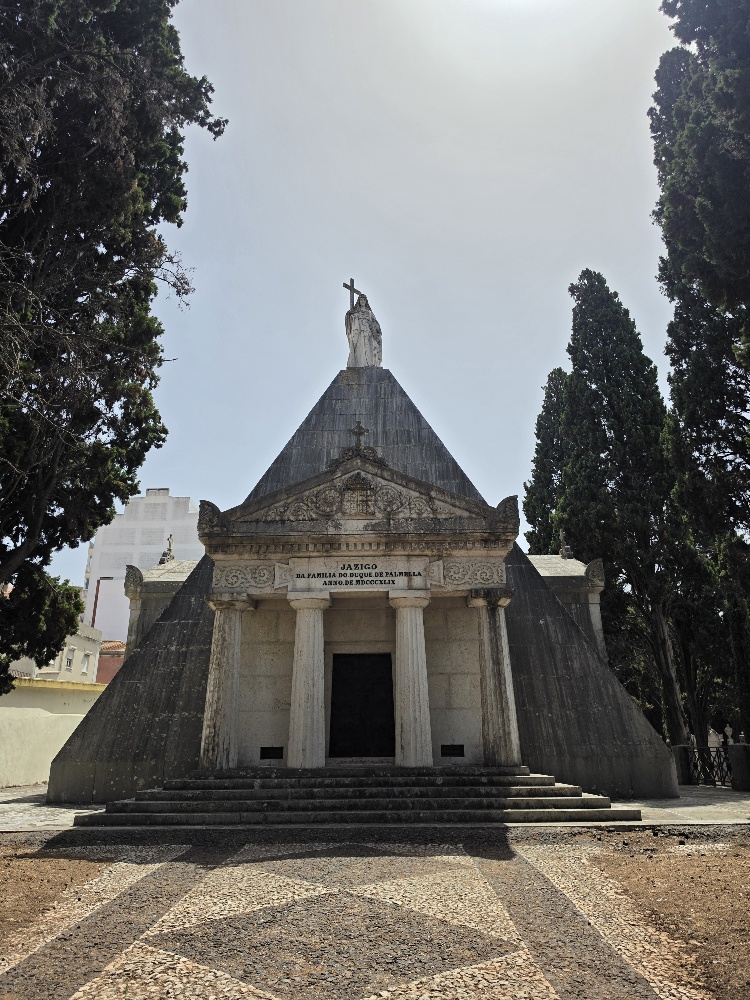 Pyramid shaped mausoleum with a statue of a person holding a cross on the top. The inscription reads Jazigo de Familia dos Duques de Palmella