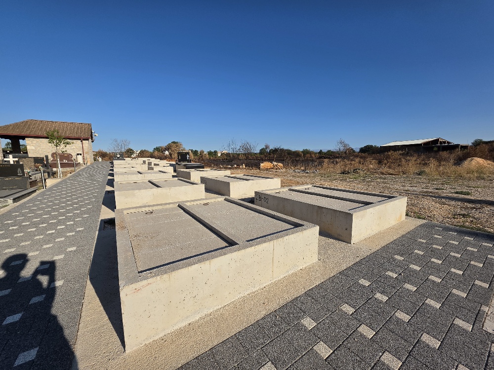 Rows of newly constructed rectangular concrete burial chambers in a cemetery, with paved walkways and an older cemetery building in the background.