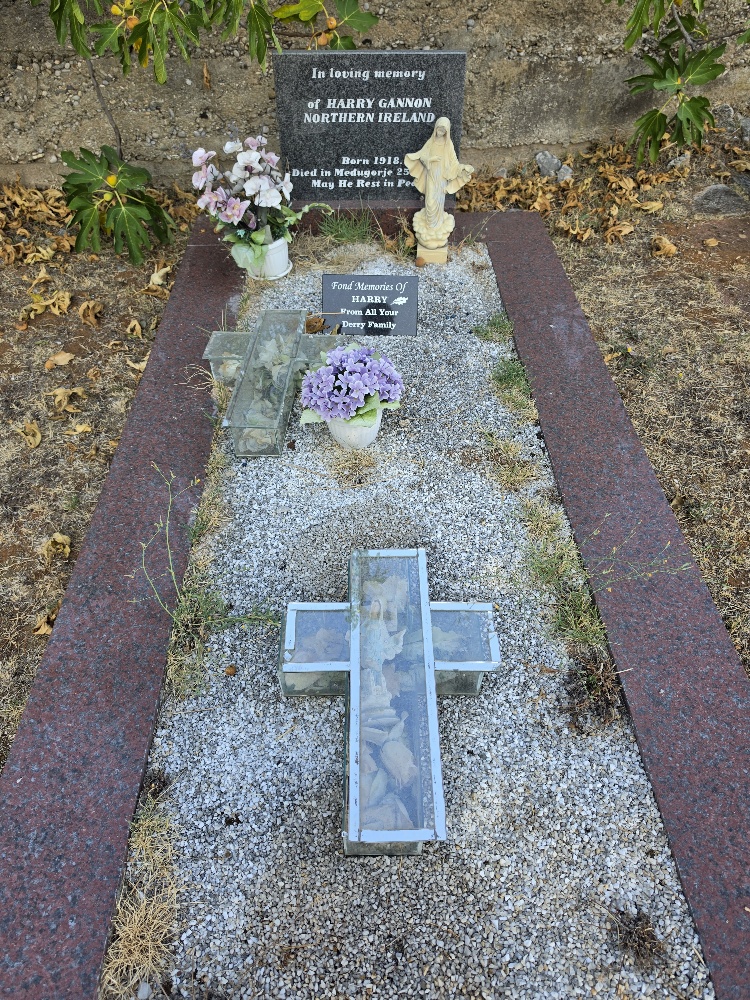 A decorated cemetery grave covered with white gravel, flowers, religious statues, and a large glass-covered cross, with a memorial plaque at the head.
