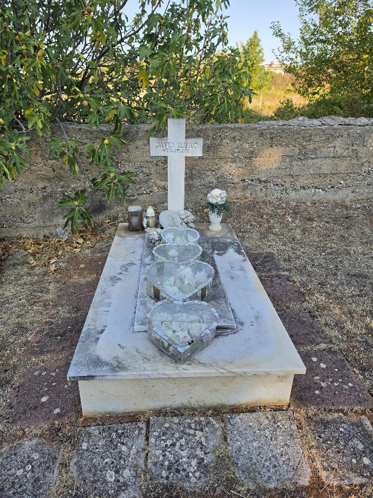 A simple white grave monument with a cross and heart-shaped glass decorations, surrounded by flowers and set against an old stone cemetery