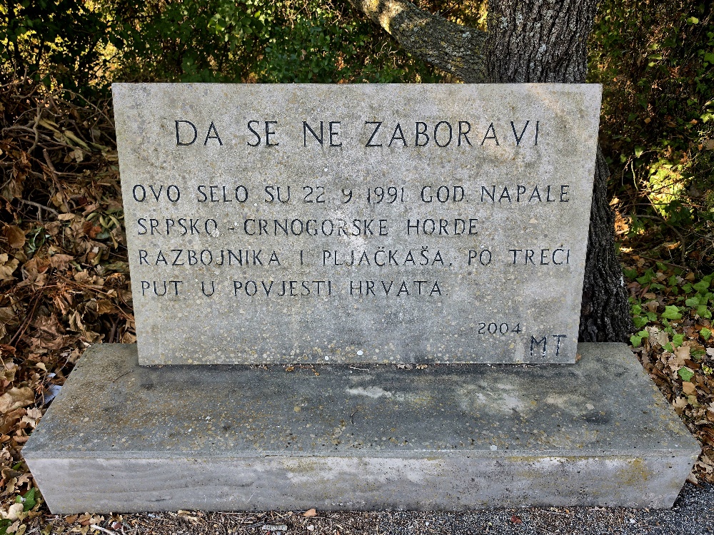 Stone memorial plaque in Croatian set against trees in Vitaljina, Konavle. The inscription commemorates an attack on the village on 22 September 1991 by Serbian-Montenegrin forces and is dated 2004. “DA SE NE ZABORAVI OVO SELO SU 22. 9. 1991. GOD. NAPALE SRPSKO-CRNOGORSKE HORDE RAZBOJNIKA I PLJAČKAŠA, PO TREĆI PUT U POVIJESTI HRVATA 2004. MT”
