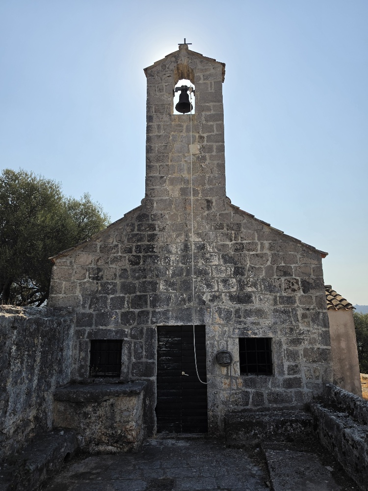Small historic stone church of Sv. Spas in Vitaljina, Croatia, with a tiled roof and arched bell gable, surrounded by the cemetery and olive trees.