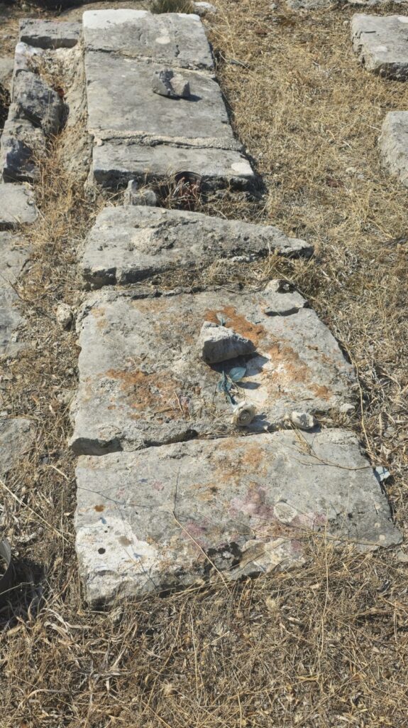 Close view of several old, weathered limestone grave slabs lying in a row in the cemetery at Sv. Spas, Vitaljina, showing rough surfaces, broken edges, lichen and signs of erosion.