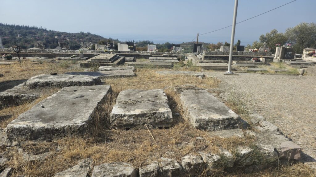 Several large, heavily weathered rectangular limestone slabs lying among grass and modern graves in the cemetery at Sv. Spas, Vitaljina; the stones are associated with the site's medieval burial ground.